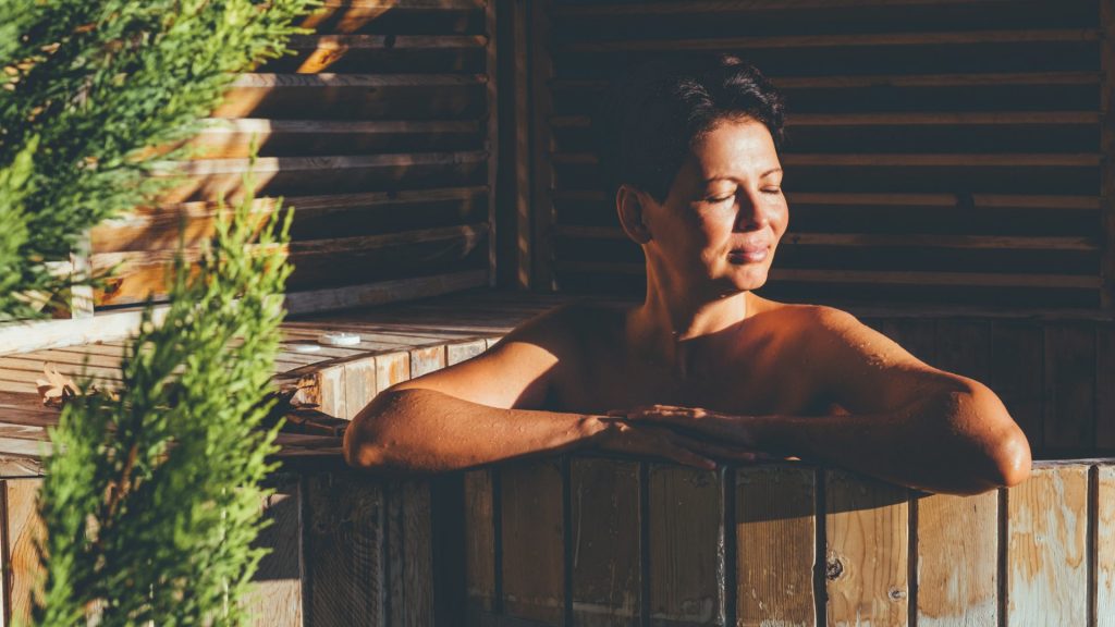 Woman sitting in a wooden hot tub with greenery next to it.