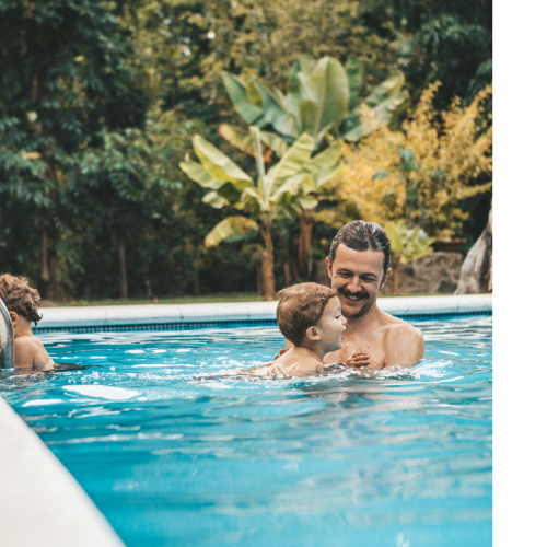 Father and young son swimming in an outdoor pool with greenery surrounding it.