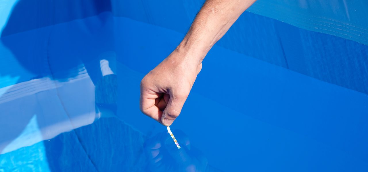 A hand reaches over the pool water to dip a test strip into it