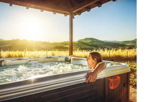 Woman sits in an outdoor hot tub with her arm hung on the edge of the spa. The sun is setting into the hills in the distance