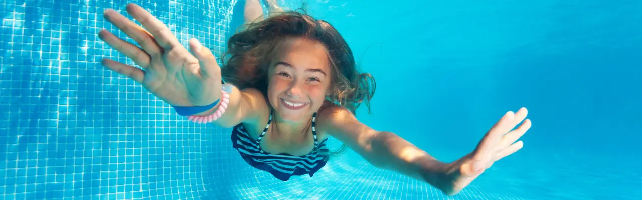 Smiling girl swimming underwater in a clear blue pool, reaching out with both hands, surrounded by a tiled pool background.