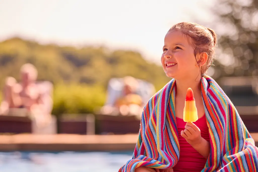 A young girl enjoys a colorful popsicle while she sits on the edge of an outdoor pool. She is wrapped in a colorful towel and smiling brightly,