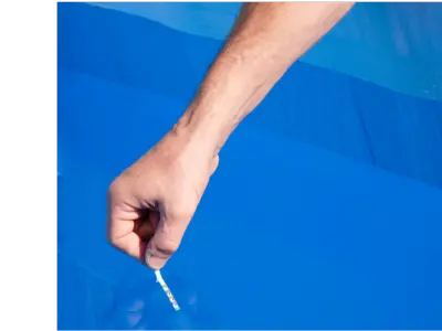 A man's hand dips a pool test strip into a vivid blue pool.