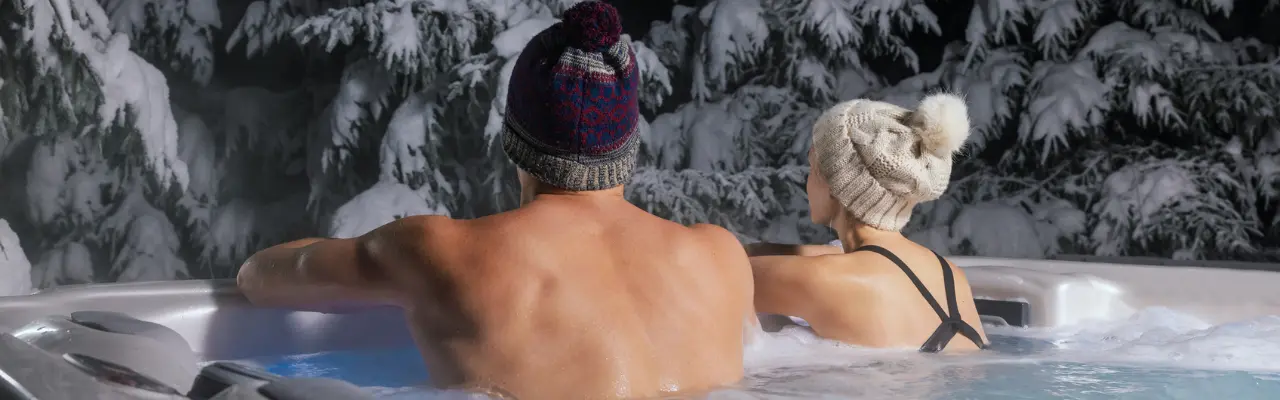 A couple sits together in their outdoor swim spa. In front of them are snow-coated trees.