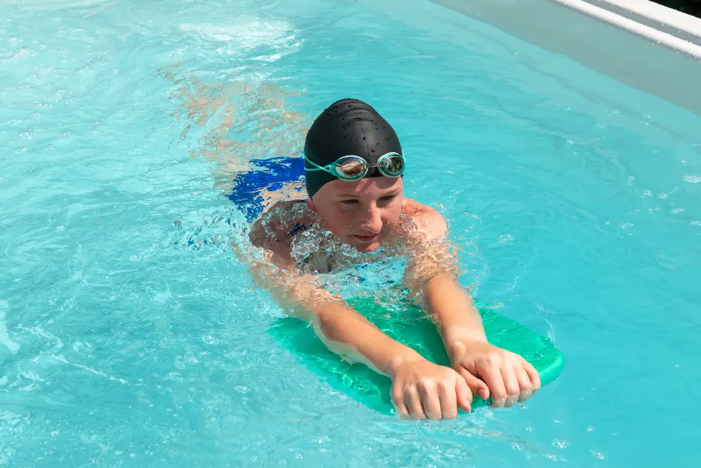 A young girl swims in a swim spa with a kickboard. She is wearing goggles and a blue swimsuit.