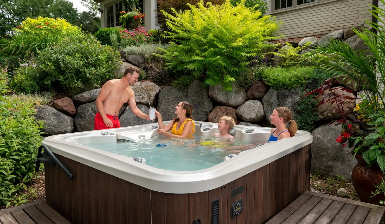 Family relaxing in an outdoor hot tub with landscaped garden surroundings, one person standing outside offering a drink
